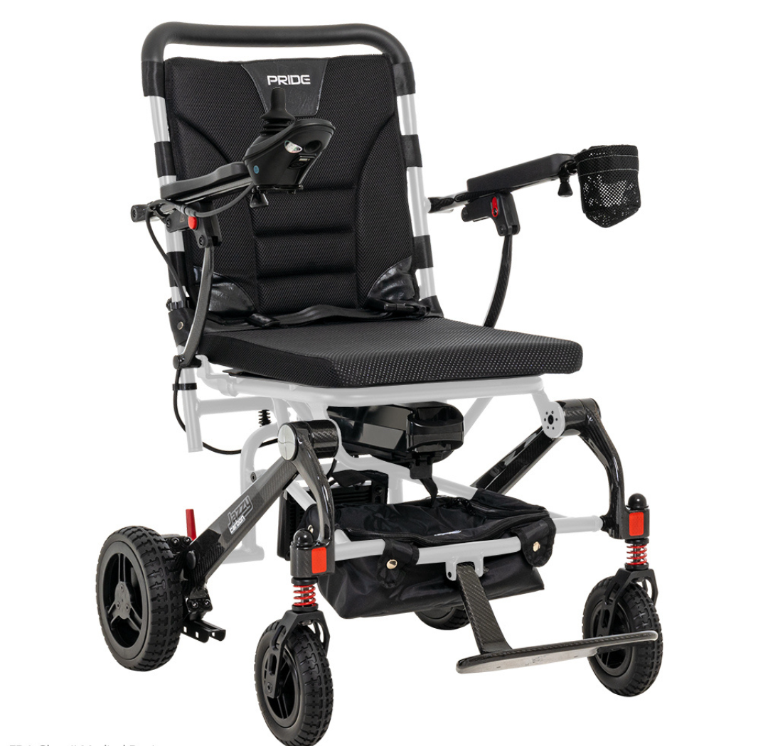 The Pride Mobility Jazzy® Carbon Ultralite Powerchair is a black and silver travel chair with cushioned seat, armrests, footrest, small front wheels, larger rear wheels, and a detachable joystick with USB charger. It includes a mesh cup holder and is labeled FDA Class II Medical Device.